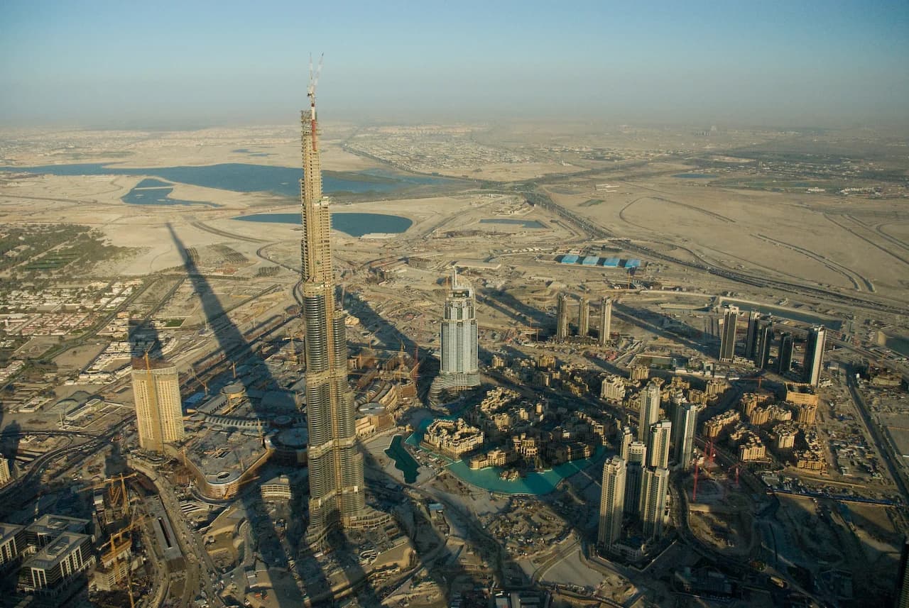 The view from At the Top, Burj Khalifa, 148th floor at sunset — the city a miniature beneath your feet, the Palm Jumeirah visible to the west, the Arabian Gulf and the desert meeting at the horizon, the curvature of the Earth just perceptible in the distance, the sense of standing on a human achievement so audacious that it provokes, simultaneously, pride in what we can build and disquiet at the cost of building it