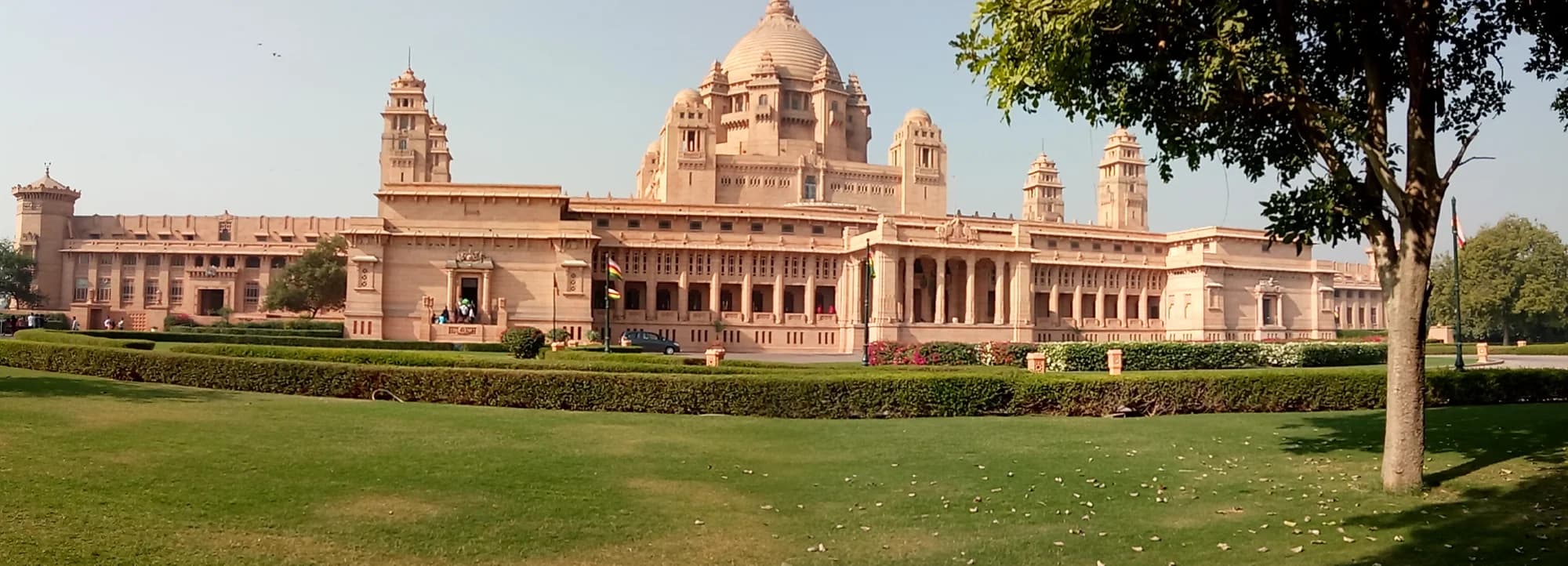 The grand entrance portico at Umaid Bhawan Palace — liveried staff in traditional Jodhpuri attire await guests at the porte-cochère, where the Maharaja's vintage Rolls-Royce and Bentley motorcars are permanently stationed as a tribute to the family's automotive passion