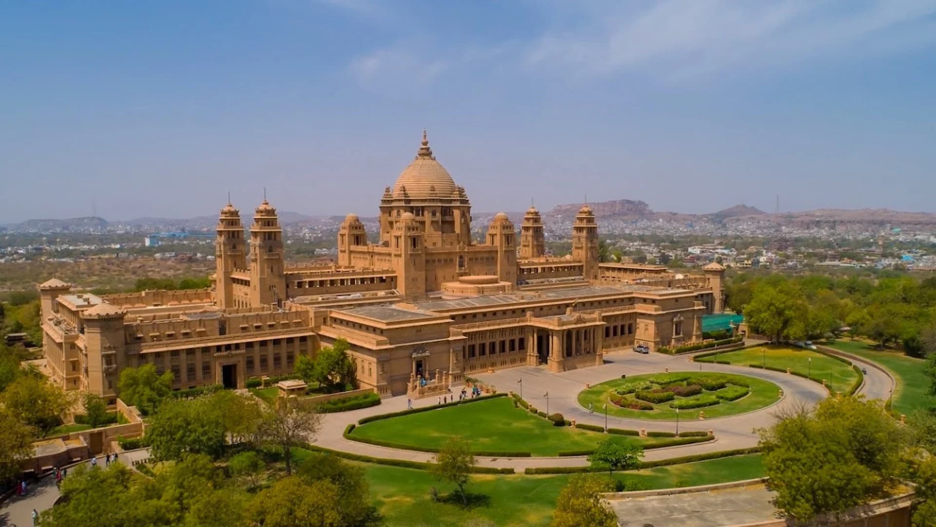 Aerial view of Umaid Bhawan Palace at sunset — 26 acres of manicured grounds spread across Chittar Hill, the crescent wings embracing formal gardens, Chittar sandstone glowing gold against the deepening desert sky