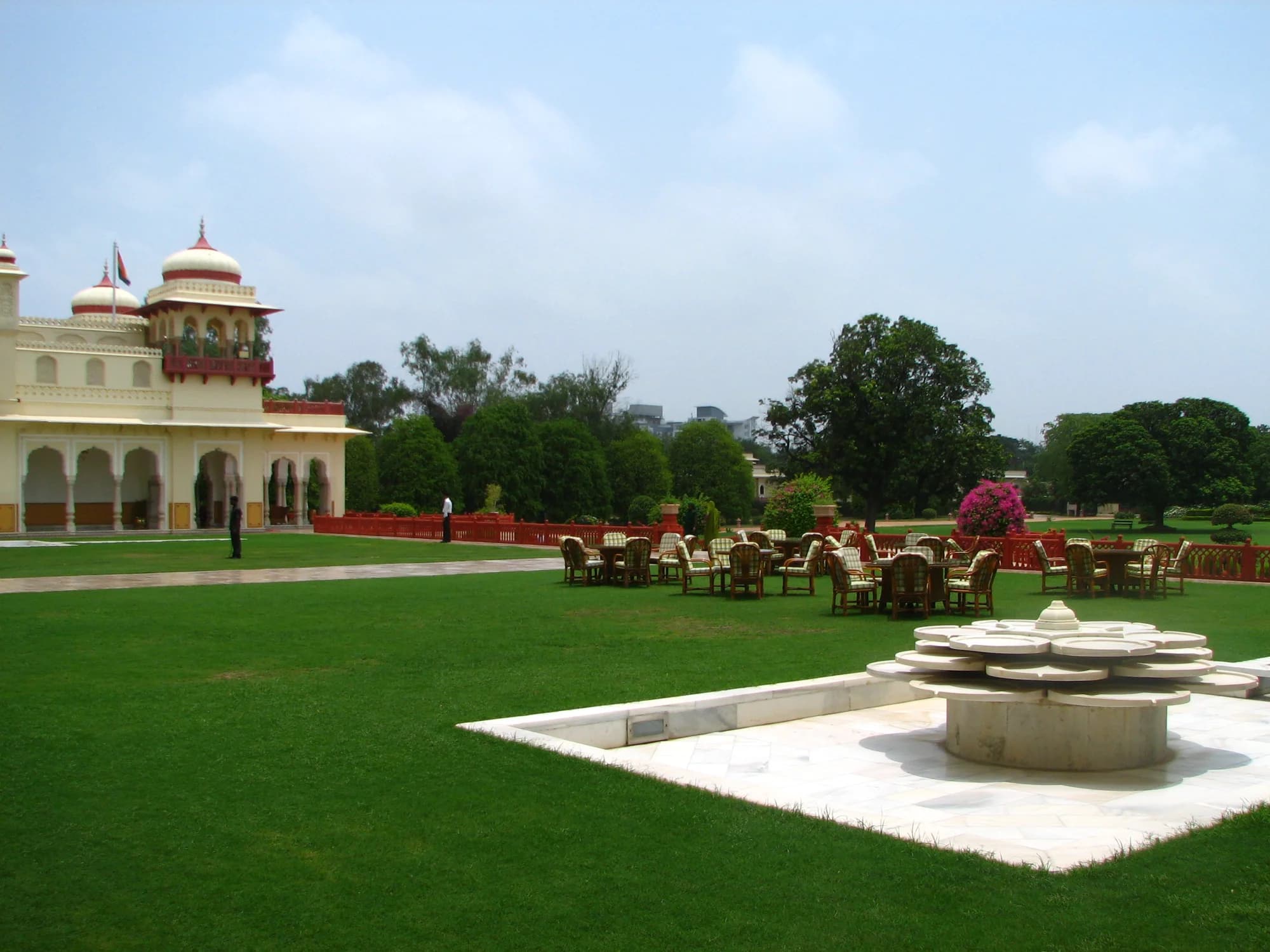 Rambagh Palace viewed from the Mughal Gardens at dawn — the ochre facade catching first light, peacocks crossing immaculate lawns, the Aravalli Hills a soft silhouette on the horizon