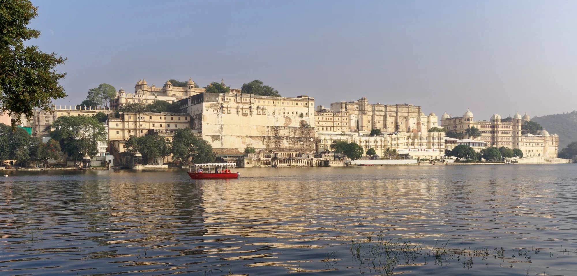 The pool terrace at Devi Garh — turquoise water set into white marble decking, the Aravalli Hills stretching to the horizon, white linen umbrellas providing the only shade under the Rajasthani sun