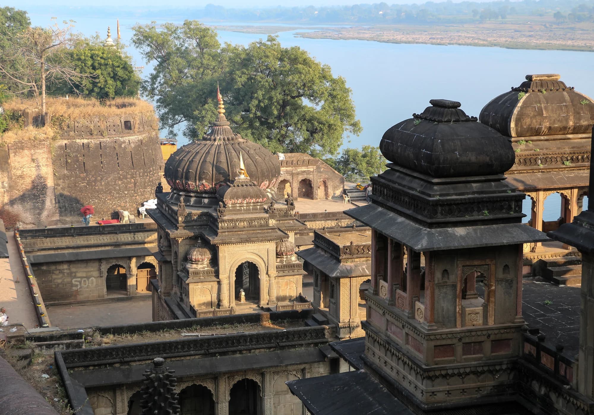 The Narmada River at sunset — pilgrims bathing at the ghats, the spires of Maheshwar's temples silhouetted against a golden sky, a boatman poling his craft across the sacred water, the chant of the evening aarti drifting across the river