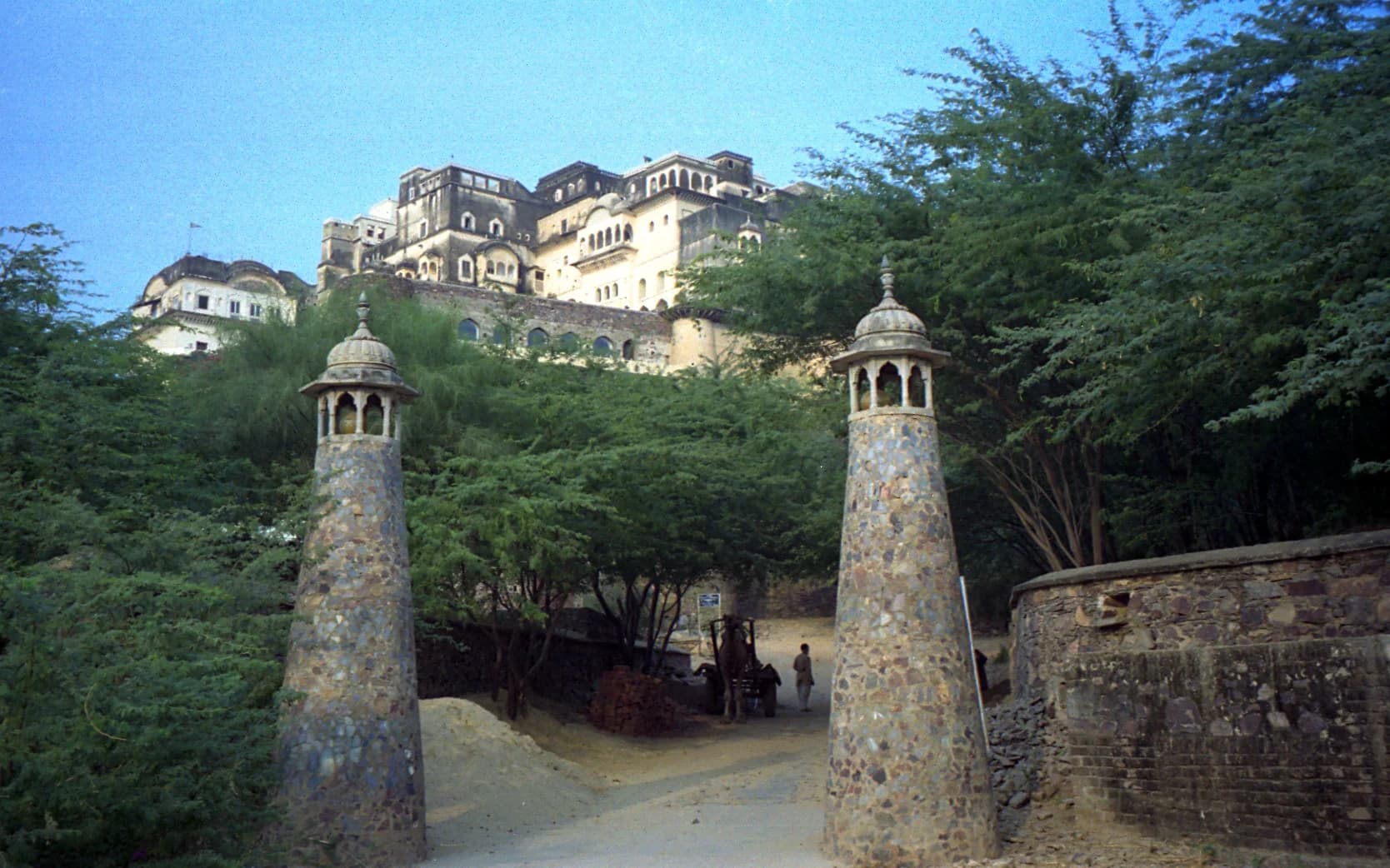 A higher-category room at Neemrana — hand-painted frescoes covering the walls, a carved four-poster bed, French windows opening onto a private balcony with a panoramic view of the Aravalli Hills