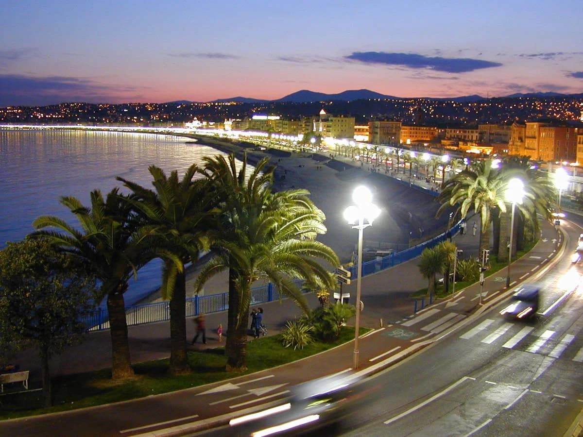 The Bay of Cannes from Le Suquet at golden hour — superyachts at anchor in the Mediterranean, the Esterel Massif blushing rose in the evening light, the Croisette a curving ribbon of palm trees and Belle Époque facades, the sky a vast canvas of gold, rose, and pale violet