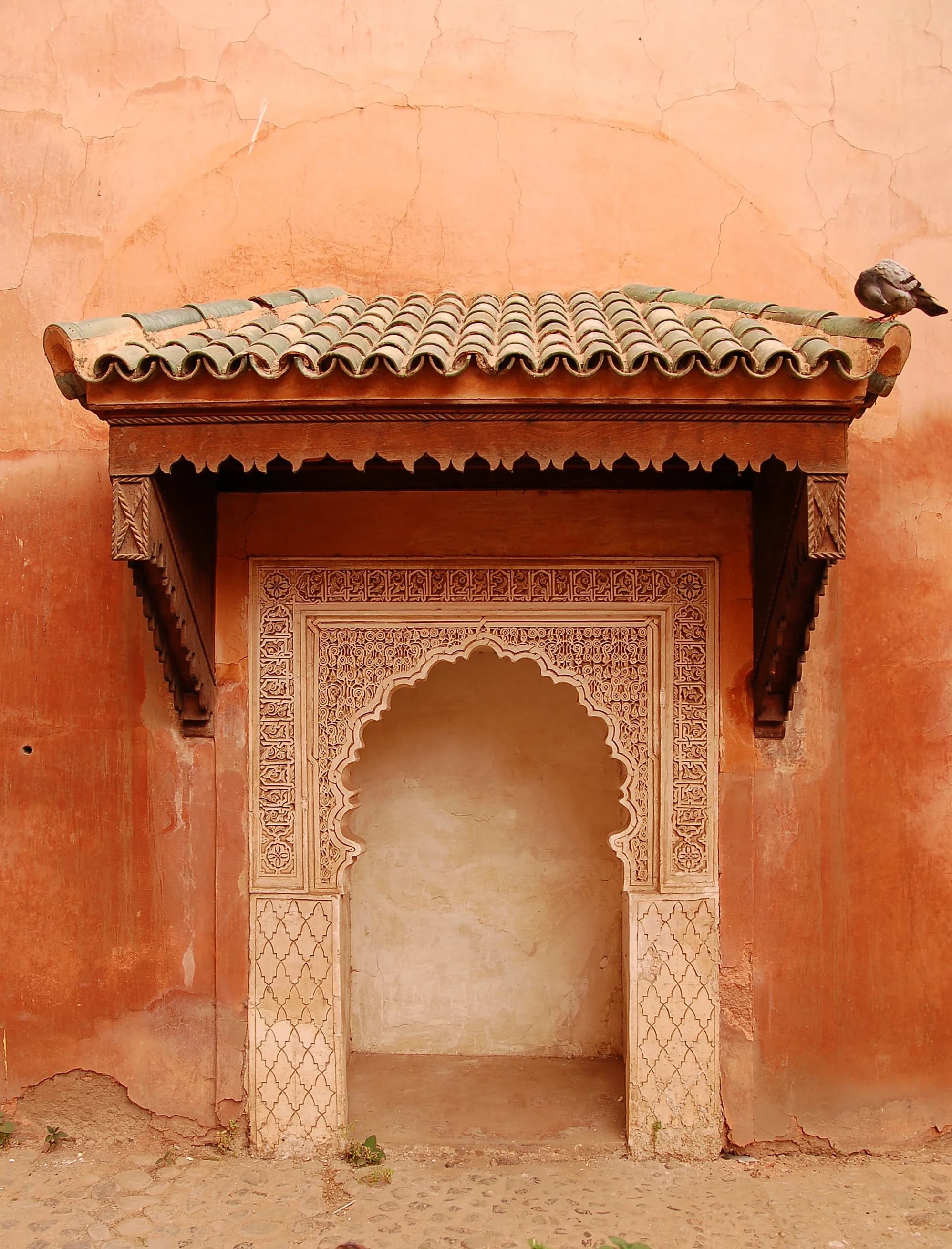 A riad courtyard at midday — the zellige tiles in patterns of green, blue, and white, the fountain at the centre filling the air with the quiet sound of water, the citrus trees casting dappled shade across the central courtyard, the intricate carved plaster of the walls and the painted cedarwood of the ceiling testifying to the centuries of craft tradition that created the Moroccan interior, the chaos of the medina a world away and a moment's walk