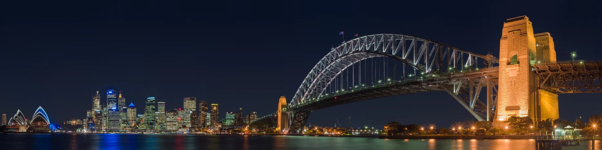 The Sydney Opera House and Harbour Bridge at dawn — Jørn Utzon's white shells glowing rose-gold in the first light, a green-and-gold ferry carving a white wake across the indigo water, the city skyline rising behind the Royal Botanic Garden