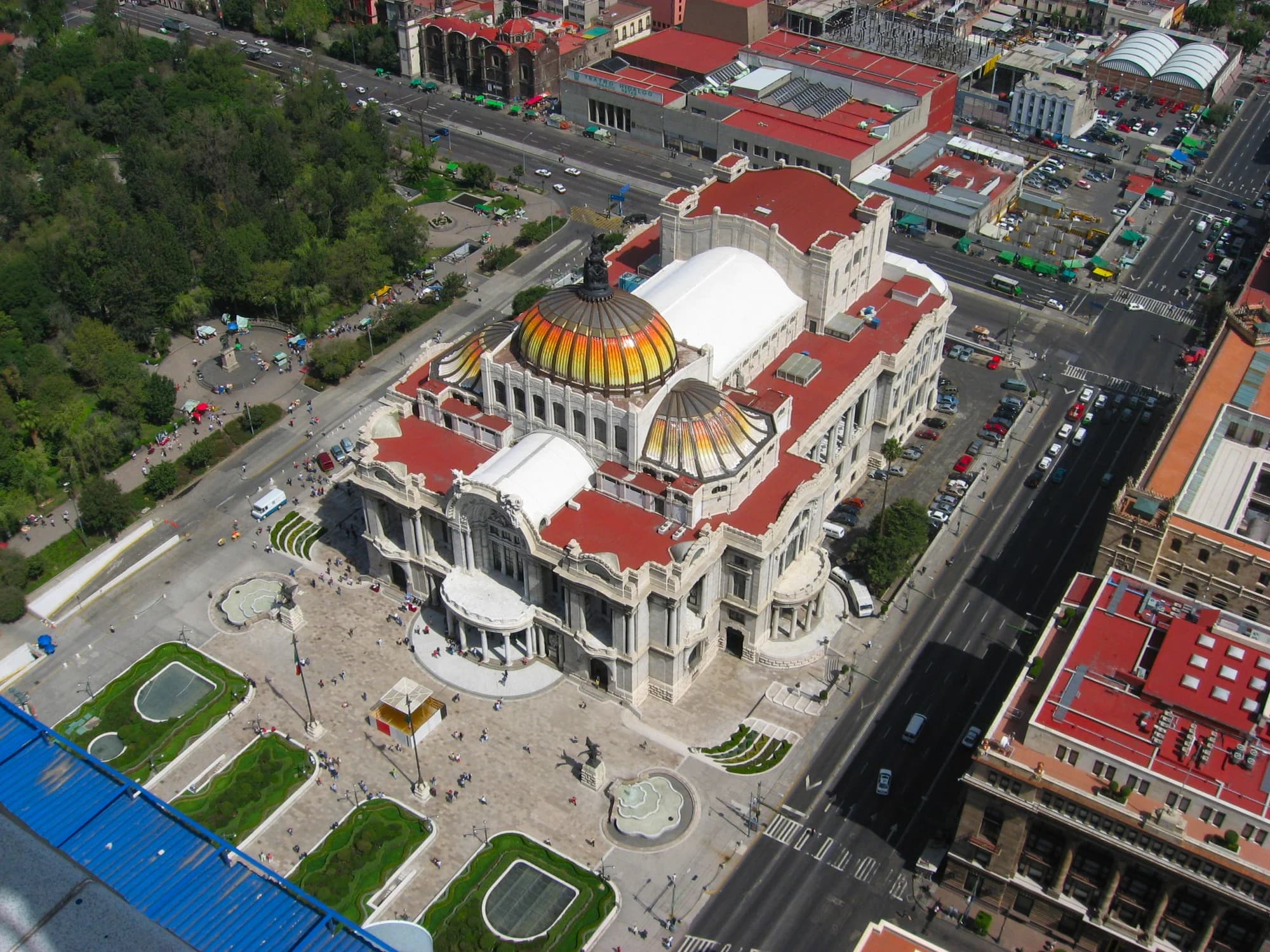 The Palacio de Bellas Artes at golden hour — the Art Nouveau and Art Deco masterpiece of Italian marble and Mexican fantasy glowing in the last light, the Torre Latinoamericana (the skyscraper that survived the 1985 earthquake) rising behind, the Alameda Central — the oldest public park in the Americas — a green carpet in the foreground, the chaotic, beautiful, impossible centre of one of the world's great cities doing what it does best: being magnificent